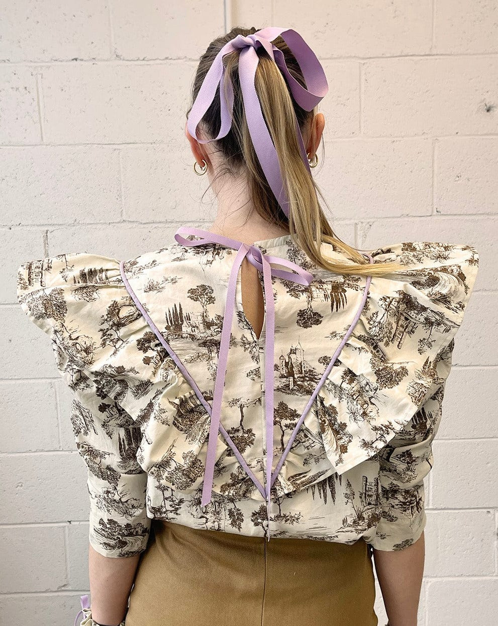 brown Toile de Jouy with bold lilac piping and matching grosgrain ribbon to really make it pop. The leg-o-mutton puff sleeves are lined with tulle for maximum volume. 100% Cotton. Slips on over head and ties in a bow at the back neck