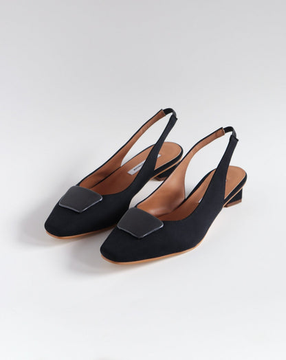 Slingback flats in a black vegan satin, made with a square toe, a unique hexagonal heel of 2.5 cm high, and an ornament piece on top that is lined with a charcoal fabric.