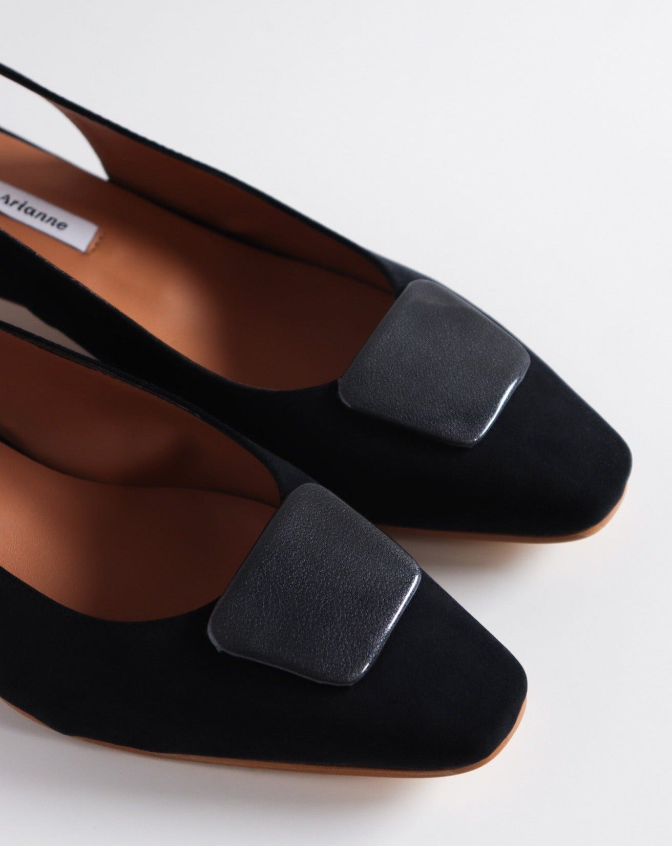 Slingback flats in a black vegan satin, made with a square toe, a unique hexagonal heel of 2.5 cm high, and an ornament piece on top that is lined with a charcoal fabric.