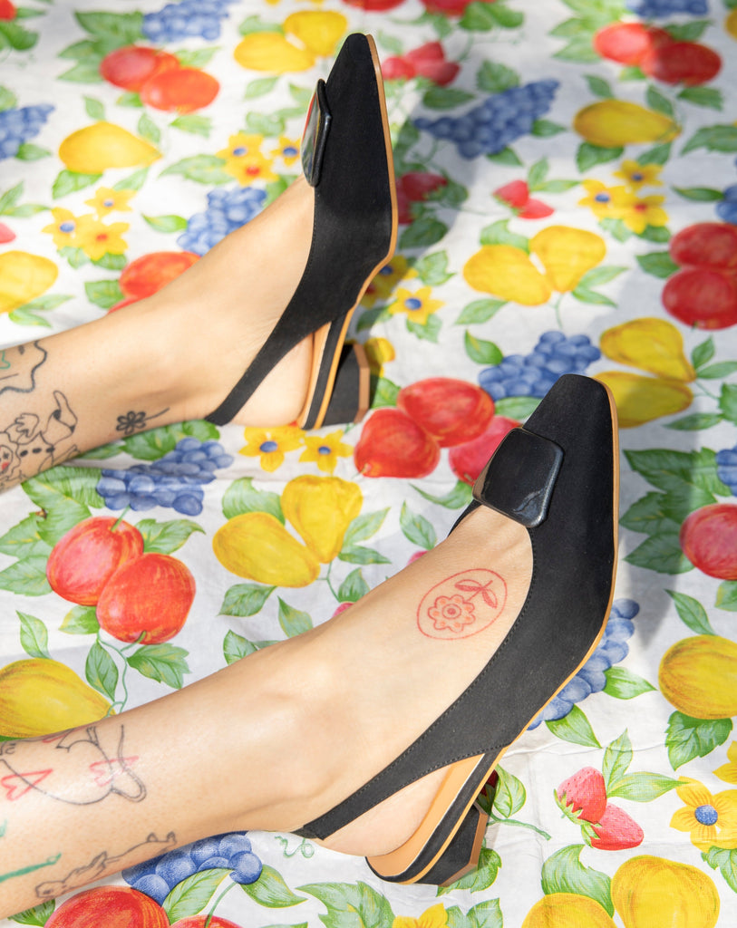 Slingback flats in a black vegan satin, made with a square toe, a unique hexagonal heel of 2.5 cm high, and an ornament piece on top that is lined with a charcoal fabric.