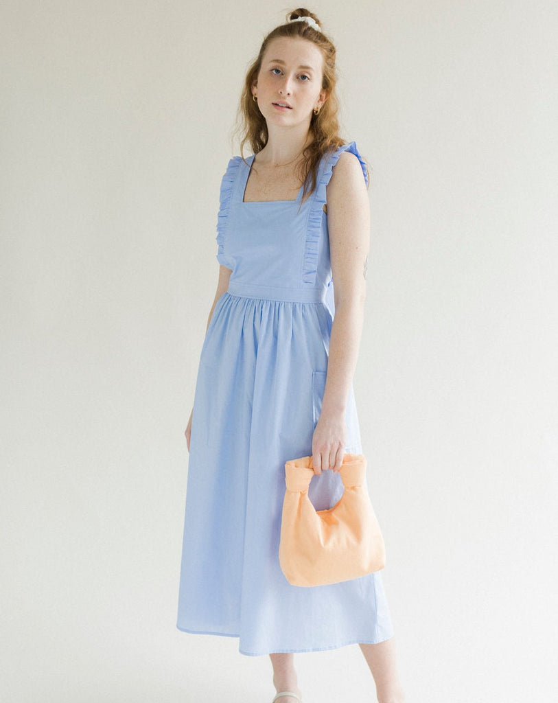 Light blue coloured apron dress with pockets, defined waist band, large straps at back to tie as bow detail, ruffled shoulder straps, and square neckline